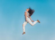 Знизу повне тіло сучасної хіпстерки-жінки в сонцезахисних окулярах у смугастій сорочці та стильних рваних джинсах, що стрибають високо на блакитне небо — стокове фото