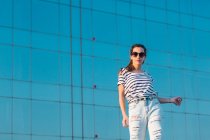 Low angle of cheerful carefree young female in sunglasses wearing casual striped t shirt and jeans enjoying sunny summer day — Stock Photo