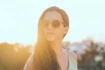 Mujer de pelo largo joven y confiada en gafas de sol de moda mirando hacia otro lado mientras pasa un día soleado de verano en el parque de la ciudad - foto de stock