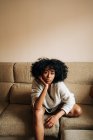 Уверенная афроамериканка с вьющимися волосами, сидящая на диване и опирающаяся на руку, глядя в камеру дома — стоковое фото
