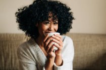 Fröhliche Afroamerikanerin mit lockigem Haar sitzt auf der Couch und trinkt Heißgetränk, während sie zu Hause in die Kamera schaut — Stockfoto