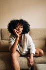 Уверенная афроамериканка с вьющимися волосами, сидящая на диване и опирающаяся на руку, глядя в камеру дома — стоковое фото