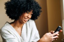 Side view of cheerful African American female sitting on soft couch in living room and browsing mobile phone at weekend at home — Stock Photo