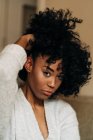 Уверенная афроамериканка с вьющимися волосами, сидящая на диване и трогающая волосы, глядя в камеру дома — стоковое фото