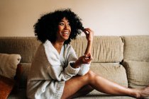 Side view of cheerful African American female with curly hair sitting on couch touching hair while looking at camera at home — Stock Photo