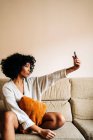 Vista laterale della femmina etnica con acconciatura afro scattare selfie sulla fotocamera dello smartphone mentre si gode il fine settimana seduto sul divano a casa — Foto stock