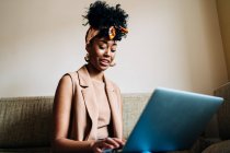 Bajo ángulo de mujer afroamericana feliz en traje elegante sentado en el sofá en casa y escribiendo en netbook mientras se trabaja en el proyecto remoto - foto de stock