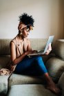Vista lateral de la mujer afroamericana en traje elegante sentado en el sofá en casa y escribiendo en netbook mientras trabaja en proyecto remoto - foto de stock