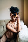 Selbstbewusste Afroamerikanerin mit modischem Haarband und Afro-Frisur sitzt auf der Couch und lehnt sich an die Hand, während sie zu Hause in die Kamera schaut — Stockfoto