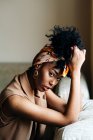 Mujer afroamericana segura con diadema de moda y con peinado afro sentado en el sofá y apoyado en la mano mientras mira la cámara en casa - foto de stock