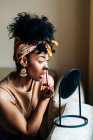 Side view of charming African American female looking in round mirror and applying powder on face while doing makeup at home — Stock Photo