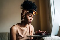 African American female in stylish outfit sitting on sofa at home and typing on netbook while working on remote project — Stock Photo