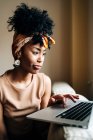 African American female in stylish outfit sitting on sofa at home and typing on netbook while working on remote project — Stock Photo