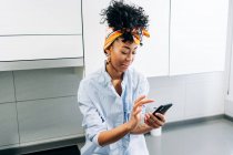 Afro-americano feminino navegar na Internet no smartphone, enquanto se situa na bancada na cozinha pela manhã — Fotografia de Stock