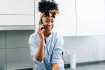African American female speaking Internet on smartphone while siting in countertop in the kitchen in morning — Stock Photo