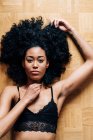 Сверху тонкая афроамериканка с вьющимися волосами, лежащими на паркетном полу и смотрящими в камеру — стоковое фото