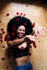 Vista superior de romántica mujer afroamericana alegre tumbada en el suelo con pétalos de rosa rojos con los ojos cerrados - foto de stock
