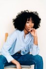 Serene black female with curly hair sitting on wooden chair against white wall and looking at camera — Stock Photo