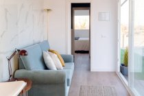 Уютный синий диван расположен в небольшой светлой комнате со стеклянной стеной при дневном свете — стоковое фото
