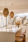 Интерьер столовой в уютной светлой кухне с плетеными бамбуковыми лампами, висящими над столом с деревянными стульями, расположенными возле окна — стоковое фото