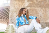 De baixo de fresco afro-americano fêmea com cabelo afro e em fones de ouvido no pescoço sentado perto de edifício urbano e olhando para a câmera — Fotografia de Stock