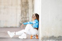 Full body side view of stylish young hipster African American female in trendy clothes and boots sitting on skateboard against concrete wall while repos on urban street — Photo de stock