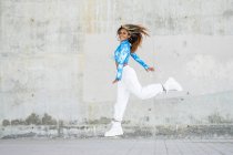 Vue latérale du corps complet de la jeune femme afro-américaine hipster optimiste en tenue informelle élégante et baskets sautant haut au-dessus du sol contre un mur en béton minable — Photo de stock