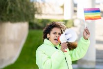 Joven activista afroamericana con bandera de arco iris LGBT en la mano gritando a un altavoz en una calle urbana borrosa - foto de stock