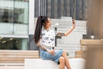 Elegante hembra asiática con el brazo tatuado descansando en el banco y tomando selfie en la calle de la ciudad - foto de stock