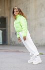 Full body side view of young African American female with dyed curly hair wearing stylish green jacket with white pants and trendy boots looking away while standing on paved urban street — Stock Photo