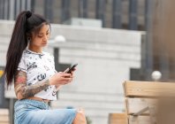 Mulher asiática elegante com braço tatuado descansando no banco e navegando telefone celular na rua da cidade — Fotografia de Stock
