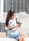 Стильна азіатська жінка з татуйованою рукою, що лежить на лавці і дивиться мобільний телефон на вулицях міста — стокове фото
