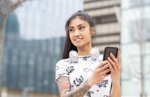 Elegante jovem mulher asiática em fones de ouvido sorrindo e tirando selfie perto do edifício moderno na rua da cidade — Fotografia de Stock