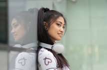Side view of pleased young Asian female with headphones smiling and looking away while leaning on glass wall on city street — Stock Photo
