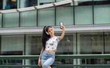 Elegante mujer asiática joven en auriculares sonriendo y tomando selfie cerca de edificio moderno en la calle de la ciudad - foto de stock