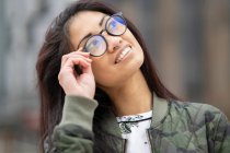 Positiva joven asiática hembra en la chaqueta de camuflaje de moda tocando gafas y mirando hacia otro lado con sonrisa en el fondo borroso de la calle de la ciudad - foto de stock