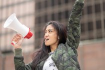 Angle bas de femme asiatique avec mégaphone levant le bras et criant pendant la manifestation dans la rue de la ville — Photo de stock