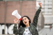 Низкий угол наклона азиатской женщины с мегафоном, поднимающей руку и кричащей во время протеста на городской улице — стоковое фото