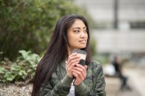 Jeune femme asiatique en tenue élégante souriant et regardant loin tout en appréciant le café pour aller le week-end dans le parc — Photo de stock