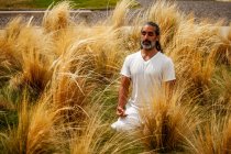 Unshaven macho hispânico em vestuário branco sentado com os olhos fechados durante a meditação entre grama dourada à luz do dia — Fotografia de Stock