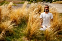 Hombre hispano sin afeitar vestido de blanco sentado con los ojos cerrados durante la meditación entre la hierba dorada a la luz del día - foto de stock