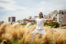 Homme ethnique barbu mature en tenue blanche étirant les bras tout en pratiquant le yoga et regardant vers l'avant contre les bâtiments urbains — Photo de stock