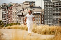 Maschio etnico barbuto di mezza età in abiti bianchi passeggiando sulla carreggiata e guardando lontano contro le case urbane — Foto stock