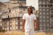 Varón barbudo de mediana edad con ropa blanca paseando por la carretera y mirando hacia las casas urbanas - foto de stock