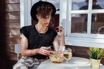 Молодой гей в модной одежде сидит со скрещенными ногами за столом с едой против бунгало — стоковое фото