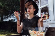 Young gay in trendy wear sitting at table with food against bungalow and white wine looking at camera — Stock Photo