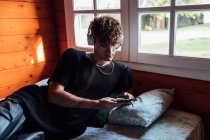 Giovane queer in cuffia wireless messaggistica di testo sul cellulare durante l'ascolto di musica sul letto in cabina — Foto stock