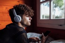 Giovane queer in cuffia wireless messaggistica di testo sul cellulare durante l'ascolto di musica sul letto in cabina — Foto stock