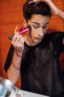 Giovane transessuale focalizzato maschio toccare i capelli mentre si applica cosmetico decorativo sul viso con applicatore contro specchio in chalet — Foto stock