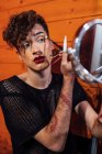 Jovem focado transexual masculino tocando cabelo ao aplicar cosméticos decorativos na sobrancelha com aplicador contra espelho em chalé — Fotografia de Stock